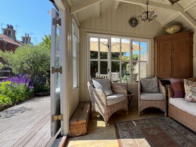 A sunroom with wicker chairs and cushions a wooden cabinet and a rug opening to a garden with flowers at Beta Cottage in Walberswick