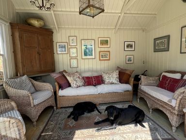 A sitting room with wicker furniture and cushions and two black dogs on a rug at Beta Cottage in Walberswick