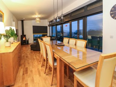 A dining area with a long wooden table and chairs next to large windows and a living area with a wood burning stove at 2 Strathtay Lodges in Aberfeldy