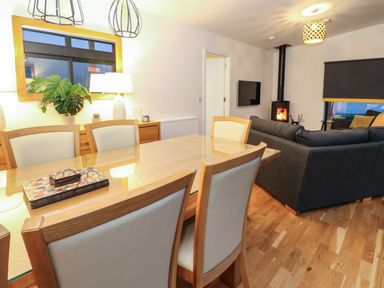 A dining area with a wooden table and chairs and a living room with a gray sofa and a wood burning stove at 2 Strathtay Lodges Aberfeldy