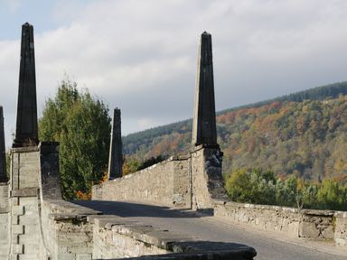 A stone bridge with obelisk-like pillars and trees with autumn foliage in the background at 2 Strathtay Lodges in Aberfeldy