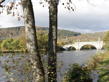 A river with a stone bridge and trees in the foreground and hills in the background at 2 Strathtay Lodges in Aberfeldy