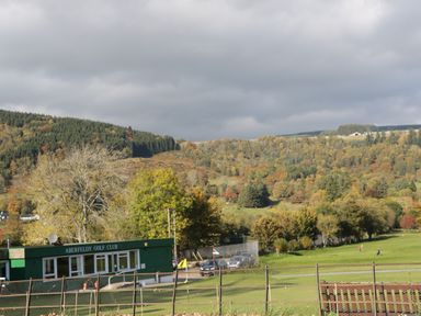 A golf course with a green club building and hills covered with trees in autumn colors at 2 Strathtay Lodges Aberfeldy