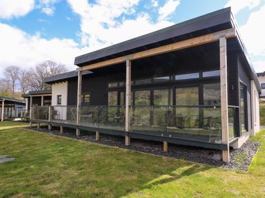 A modern single story house with a wooden porch and glass railing overlooking a grassy yard at 2 Strathtay Lodges in Aberfeldy