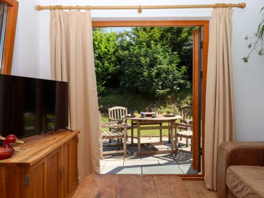A living room with a TV on a wooden cabinet and an open door leading to a patio with a wooden table and chairs at South Holm in Moreleigh