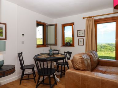 A dining area with a round black table set with placemats and glasses and black chairs beside a brown leather sofa with windows and a glass door showing a countryside view at South Holm in Moreleigh