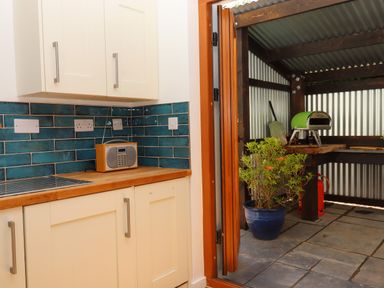A kitchen corner with white cabinets blue tile backsplash and a radio next to an open door leading to a covered outdoor area with a plant a small oven and tiled floor at South Holm in Moreleigh