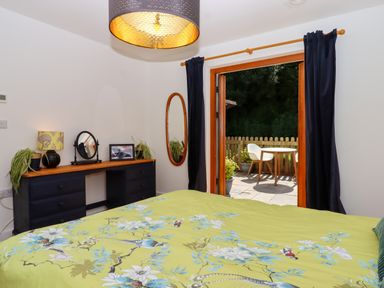 A bedroom with a green floral bedspread a dresser with plants and a mirror and an open door leading to a patio with chairs at South Holm in Moreleigh
