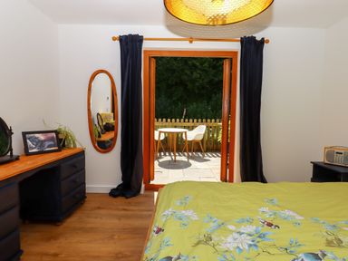 A bedroom with a bed covered in a green floral duvet a wooden dresser with a mirror and photo frame and an open door leading to a patio with a small table and two white chairs at South Holm in Moreleigh
