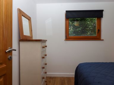 A bedroom with a wooden dresser and mirror next to a window with a black blind and part of a bed with a blue quilt at South Holm in Moreleigh