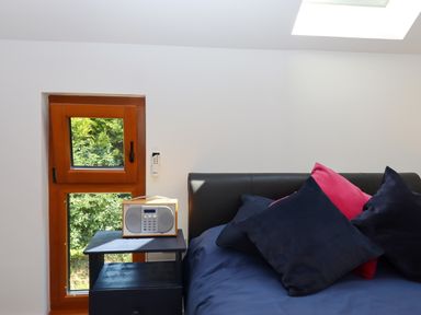 A bedroom corner with a bed with blue and pink pillows next to a wooden window and a small black bedside table with a radio on it at South Holm in Moreleigh
