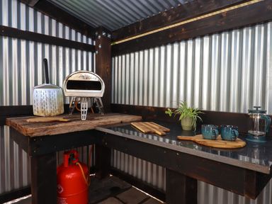 A small outdoor kitchen with a wood-fired oven pizza peel cutting boards two mugs a plant and a gas canister at South Holm in Moreleigh