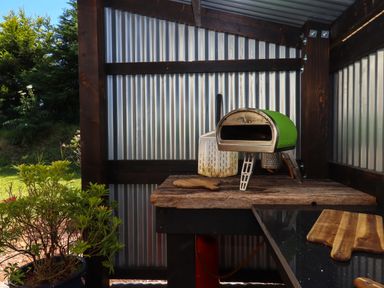 An outdoor cooking area with a green pizza oven wooden cutting boards and a plant in a pot at South Holm in Moreleigh