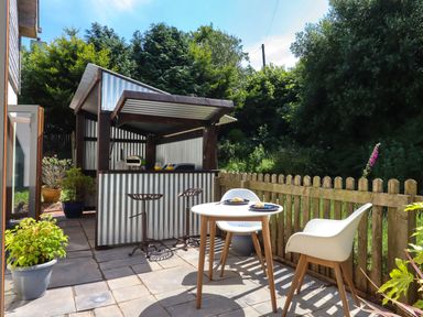 An outdoor patio area with a small round table and two chairs near a picket fence and a covered metal bar with stools at South Holm in Moreleigh