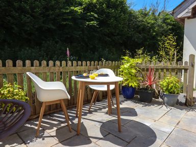 An outdoor patio with a small round table set for two and white chairs next to a wooden fence with potted plants at South Holm in Moreleigh