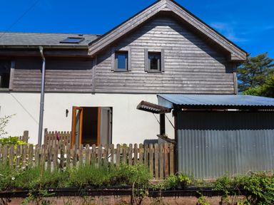 A wooden fence, garden with plants, shed with corrugated metal roof, and a house with wooden siding and an open door at South Holm in Moreleigh
