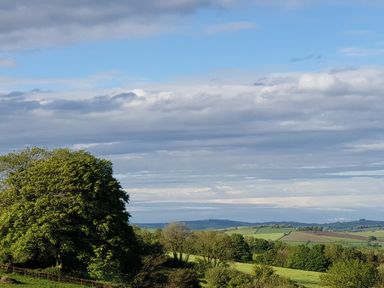 A rural landscape with green fields trees and hills under a partly cloudy sky at South Holm in Moreleigh