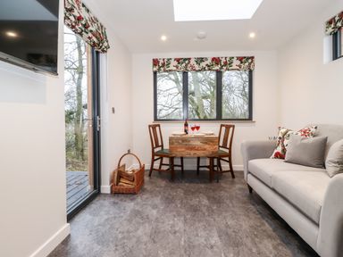 A small dining area with a wooden table and two chairs next to a window with floral curtains and a gray sofa with cushions at Herdwick Cabin in Hutton John near Penruddock