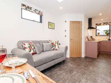 A living room with a grey sofa and floral cushions next to a kitchen with pink cabinets at Herdwick Cabin in Hutton John near Penruddock