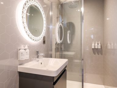 A bathroom with a sink a mirror with lights a shower area and mounted soap dispensers at Herdwick Cabin in Hutton John near Penruddock