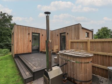 A wooden deck with two chairs and a hot tub outside a wooden cabin at Herdwick Cabin in Hutton John near Penruddock