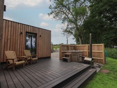 An outdoor wooden deck with two wooden chairs a small table a door a wooden hot tub and a wood stove surrounded by a wooden fence at Herdwick Cabin in Hutton John near Penruddock
