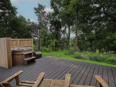 A wooden deck with two chairs and a hot tub near trees and grass at Herdwick Cabin in Hutton John near Penruddock
