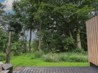 An outdoor view of trees and grass next to a wooden deck and a metal chimney at Herdwick Cabin in Hutton John near Penruddock