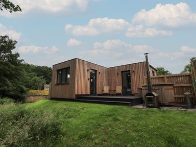 A wooden cabin with two chairs and a table on a deck and a hot tub on grass at Herdwick Cabin in Hutton John near Penruddock