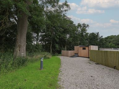 A gravel pathway with wooden fencing and a wooden structure next to green grass and trees at Herdwick Cabin in Hutton John near Penruddock