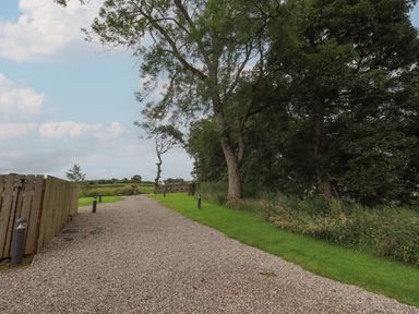 A gravel pathway bordered by grass and trees with a wooden fence on the left and countryside in the background at Herdwick Cabin in Hutton John near Penruddock