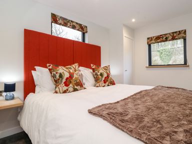 A bedroom with a bed with red headboard pillows and blanket with windows in the background at The Gardener's Retreat Hutton John near Penruddock