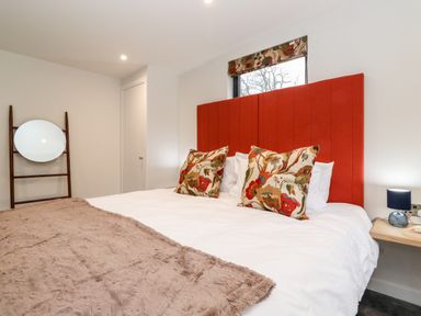 A bedroom with a red headboard two floral pillows a round mirror on a wooden ladder and a small lamp on a bedside table at The Gardener's Retreat in Hutton John near Penruddock
