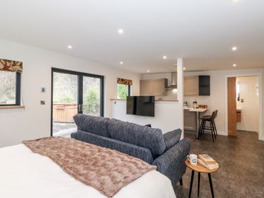 An open-plan room with a bed, sofa, small table with books and a mug, kitchen area with stools, and glass doors leading outside at The Gardener's Retreat in Hutton John near Penruddock