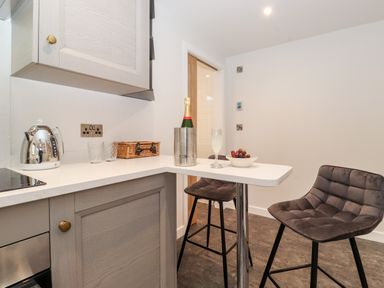 A kitchen corner with a small breakfast bar two stools a kettle glasses a wicker basket a champagne bottle in an ice bucket a glass with a drink and a bowl of grapes at The Gardener's Retreat in Hutton John near Penruddock