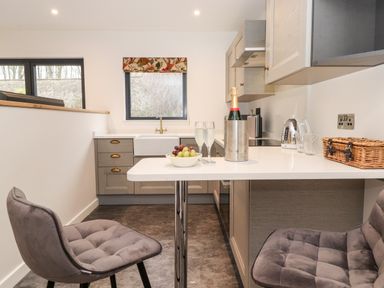 A kitchen area with two grey chairs and a white countertop with a bottle in a cooler and two glasses at The Gardener's Retreat in Hutton John near Penruddock