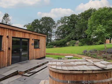 A wooden cabin with glass doors next to a covered wooden hot tub and a garden with grass trees and patio furniture at The Gardener's Retreat in Hutton John near Penruddock