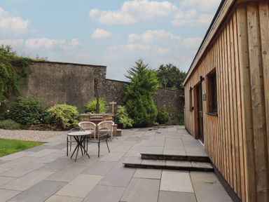 An outdoor patio with a wooden building on the right a table with chairs and plants against a stone wall at The Gardener's Retreat in Hutton John near Penruddock