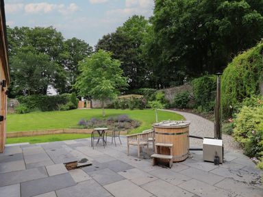 A garden with a paved patio area featuring a wooden hot tub and chairs overlooking a grassy area with trees and a gravel path at The Gardener's Retreat in Hutton John near Penruddock