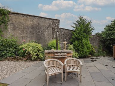 Two wooden chairs in front of a wooden hot tub on a paved patio with plants and a stone wall in the background at The Gardener's Retreat in Hutton John near Penruddock