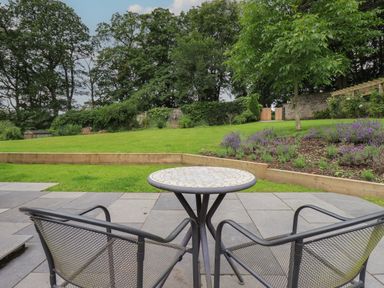 A patio with a round table and two metal mesh chairs overlooking a lawn and garden at The Gardener's Retreat in Hutton John near Penruddock