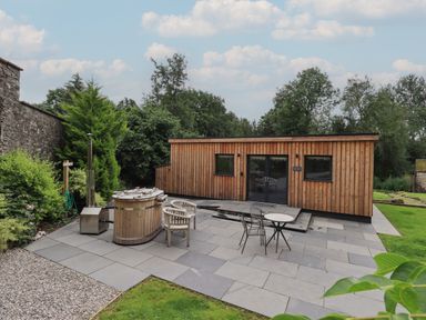 A wooden cabin with outdoor seating and a hot tub on a stone patio surrounded by grass and trees at The Gardener's Retreat in Hutton John near Penruddock