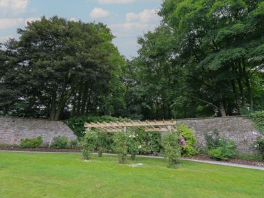A garden with a wooden pergola and green bushes in front of a stone wall and trees at The Gardener's Retreat in Hutton John near Penruddock