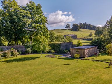 A garden with grass trees a wooden shed and a stone wall with hills in the background at The Gardener's Retreat in Hutton John near Penruddock