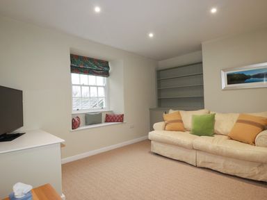 A living room with a cream sofa with cushions a window seat with cushions a television on a white unit and a built in shelving unit at Tretoil Farm House in Bodmin