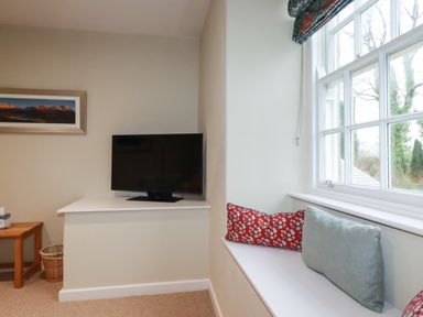A room corner with a mounted television a wooden side table with a tissue box a wicker basket and a window seat with cushions at Tretoil Farm House in Bodmin