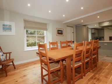 A dining room with a wooden table and eight chairs next to a window and kitchen at Tretoil Farm House in Bodmin