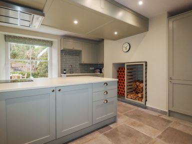 A kitchen with grey cabinets and tiled floor a window with a view of outdoor seating and a wall clock at Tretoil Farm House in Bodmin