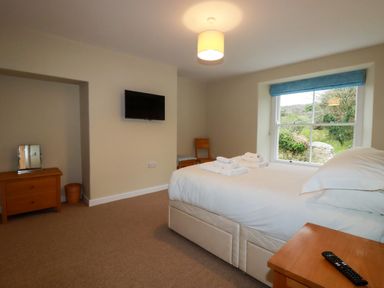 A bedroom with a bed, folded towels, a window, a wooden chair, a small dresser with a mirror, and a wall-mounted television at Tretoil Farm House in Bodmin