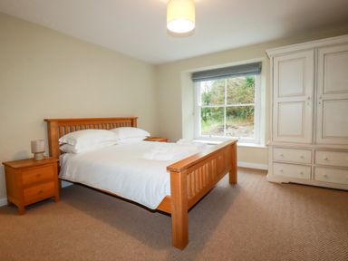 A bedroom with a wooden double bed white bedding wooden bedside tables a lamp a large white wardrobe and a window at Tretoil Farm House in Bodmin